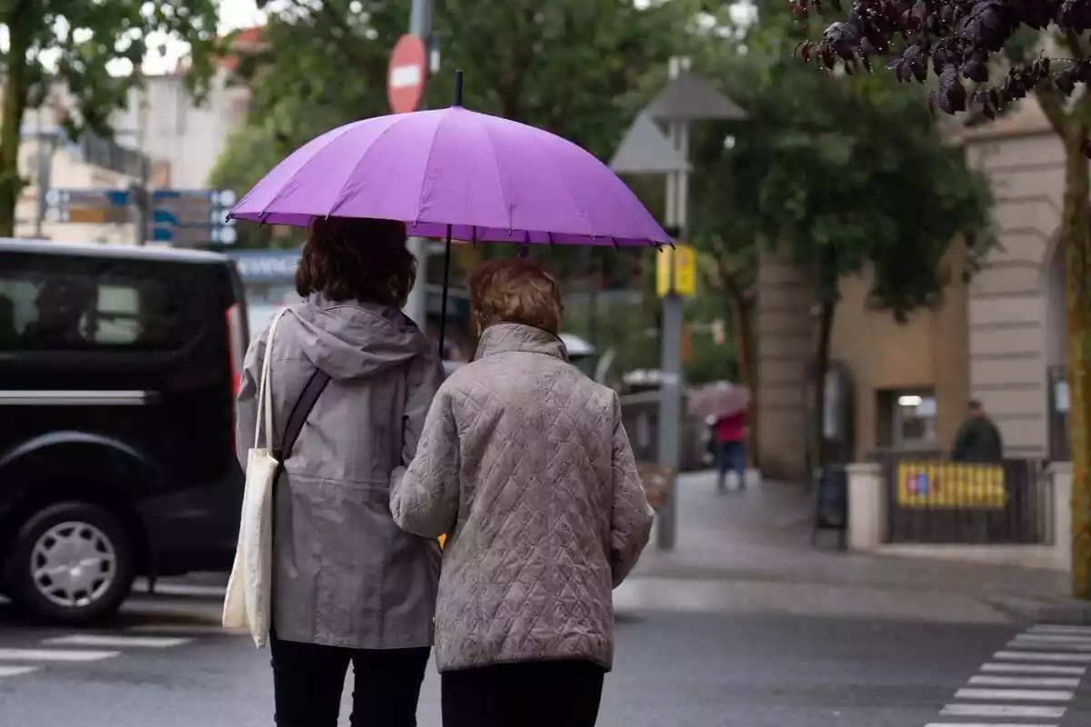 Dos mujeres de espalda caminando por la calle con chaqueta y un paraguas lila